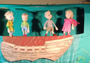 Scena w zielonym kolorze. W dolnej części zawieszony fragment łodzi. W górnej kukiełki trzech chłopców i dziewczynki. Wygląda tak, jakby wyruszali w morską podróż.